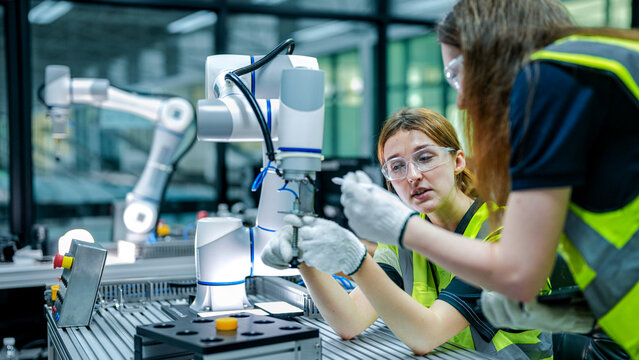 Two female robotics engineers in an R&D lab troubleshoot an AI cobot. One inspects the arm's hardware while the other reviews diagnostic data for the system. - Powered by Adobe