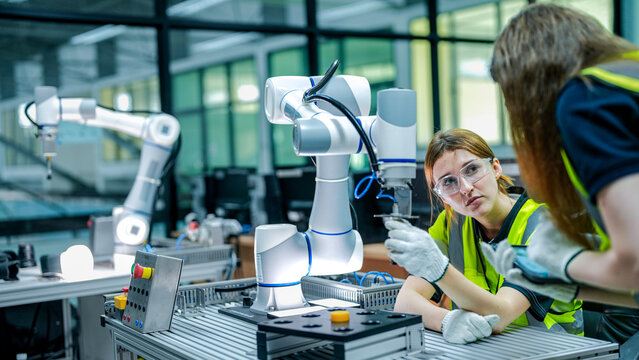 Two female robotics engineers in an R&D lab troubleshoot an AI cobot. One inspects the arm's hardware while the other reviews diagnostic data for the system.