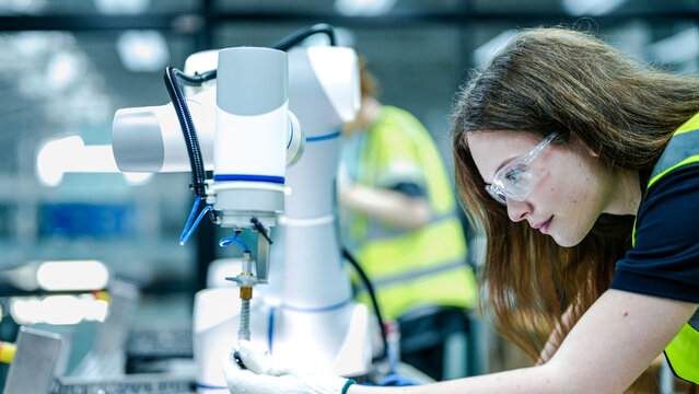 Two female robotics engineers in an R and D lab troubleshoot an artificial intelligence cobot. They are inspecting the robotic system hardware, sensors, and programming.
