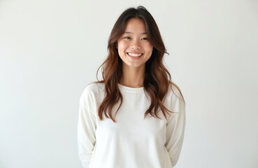 Obraz premium Young Asian woman smiles happily. She wears a simple white shirt and has long brown hair. Her portrait is taken in a studio against a plain background, looking directly at the viewer.