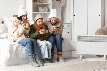 Frozen family with lack of heating drinking hot tea near electric convector heater in bedroom
