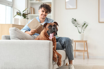 Handsome young man with book and Boxer dog sitting on sofa at home