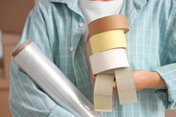 Young woman with adhesive tape rolls and stretch wrap in room on moving day, closeup