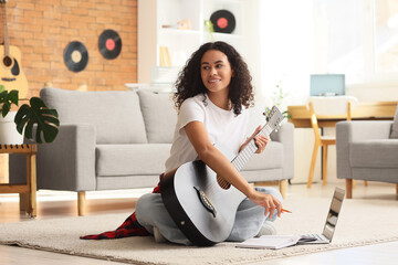 Pretty young African-American woman with laptop, notebook and acoustic guitar sitting on floor in living room