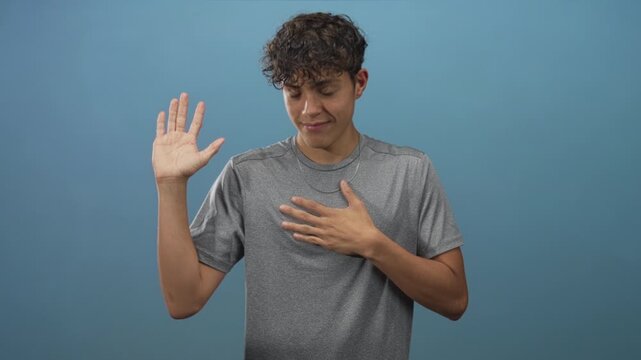 Man raising one hand while placing other hand on chest in studio against blue backdrop, wearing gray tshirt and necklace, solemn expression; sincerity.