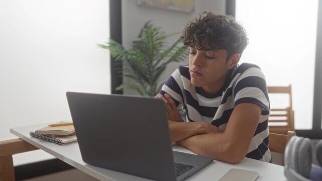 Teen boy in striped shirt with folded arms stares at laptop on desk with notebooks and headphones in building; boredom contemplation focus.