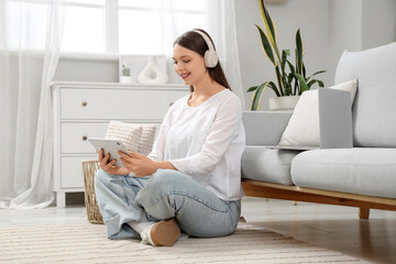 Obraz premium Beautiful young woman in headphones with modern tablet computer sitting on carpet in living room