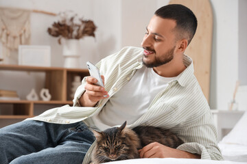 Handsome young man with mobile phone and cute cat sitting on bed in bedroom