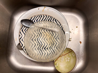 A stack of dirty dishes, including ceramic plates with patterns, a clear glass bowl, and metal spoons, sitting in a kitchen sink waiting to be washed.