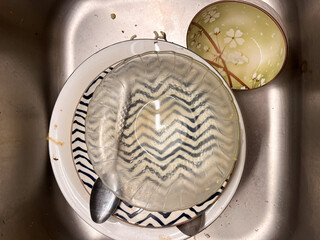 A stack of dirty dishes, including ceramic plates with patterns, a clear glass bowl, and metal spoons, sitting in a kitchen sink waiting to be washed.