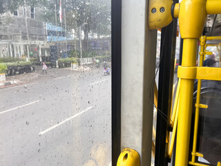 A POV (point of view) shot from inside a public bus on a rainy day. The image features close-up water droplets on a window pane, creating a soft bokeh effect of the city street and sidewalk outside.