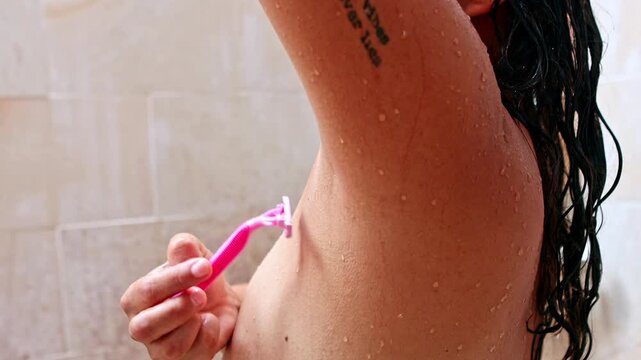 Tattooed woman shaving armpit with razor in the shower