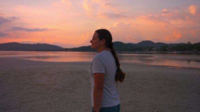 Rear Tracking Shot of Woman Walking Slowly across Shallow Tidal Flat with Golden Sunset Light Reflecting on Calm Water under Heavy Clouds, Emotional Balance Concept