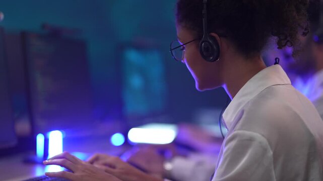 Rear view of a female call center agent with curly hair wearing a headset. She is sitting in a row with colleagues in a dark, blue-lit office. The scene represents a modern customer support team worki