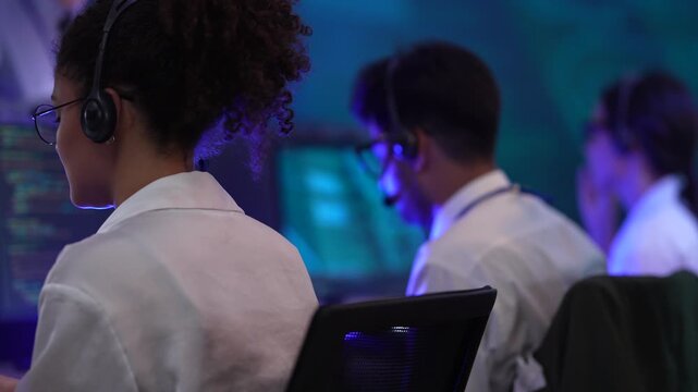 Rear view of a female call center agent with curly hair wearing a headset. She is sitting in a row with colleagues in a dark, blue-lit office. The scene represents a modern customer support team worki