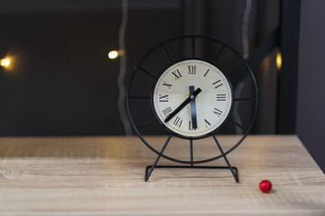 Round black metal desk clock with roman numerals on a wooden table, against a dark background with...
