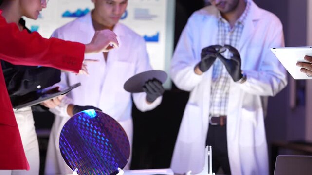 A diverse group of scientists and business professionals standing around a large silicon wafer. A male scientist in a white coat and black gloves presents the shiny disk to a woman in a red suit holdi