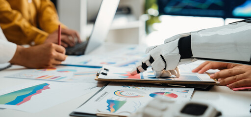 A robotic arm assists professionals in analyzing financial data within a contemporary office space, exemplifying the fusion of technology and business for enhanced productivity.Noogenesis