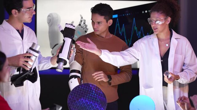 Diverse group of scientists and engineers examining a white robotic prosthetic arm and a large silicon wafer. A smiling woman in safety glasses and a man in a white lab coat are discussing the innovat