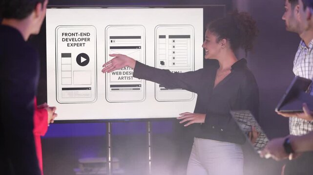 A smiling woman with curly hair discussing mobile app wireframes on a large presentation screen with a male colleague holding a laptop. The screen displays sketches for "Front-End Developer Expert" an