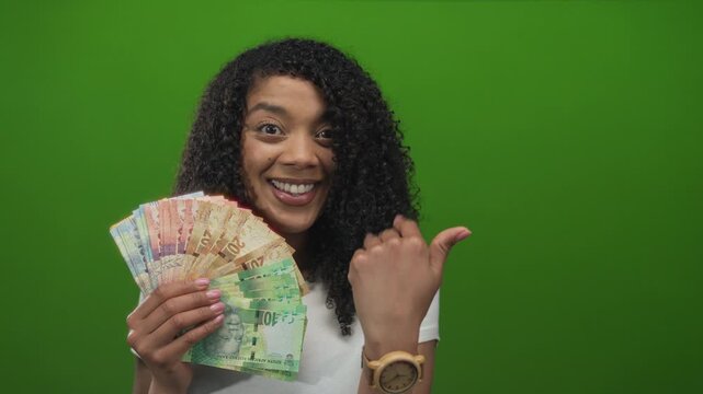 Woman holds south african rand banknotes, smiles brightly and shows open palm in studio; financial success.
