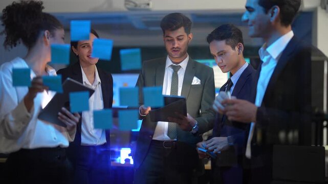 A young businessman in a suit writing on a glass wall with a green marker during a brainstorming session. A woman with curly hair stands nearby looking at blue sticky notes. Colleagues in the backgrou