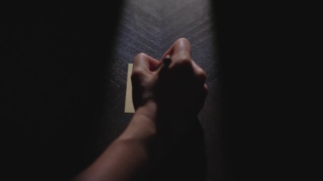 Slow motion close up footage of a hand writing out "" on a paper note and placing it on a table. Shot in dramatic lighting for an impressive title or transition.