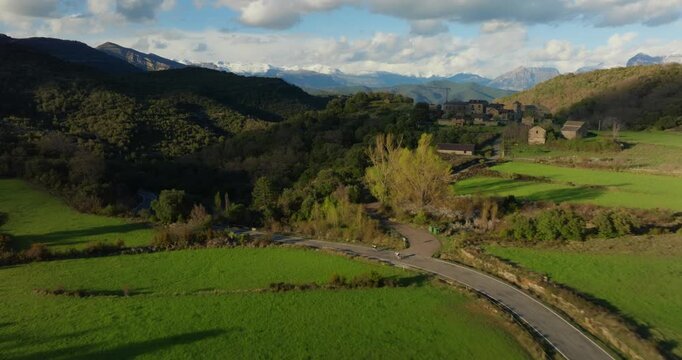 Drone tracking shot follows cyclists riding along HU‑641 mountain road linking Mirador de Fuebla to A&iacute;nsa in Huesca, Aragon, Spain, above bright green valley farmland and distant Pyrenean peaks