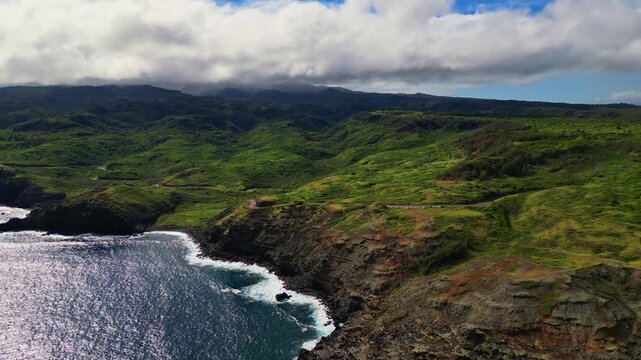An aerial shot of Maui's rugged coastline with green hills cascading to the ocean, white waves crashing against rocks under a sky with fluffy clouds.