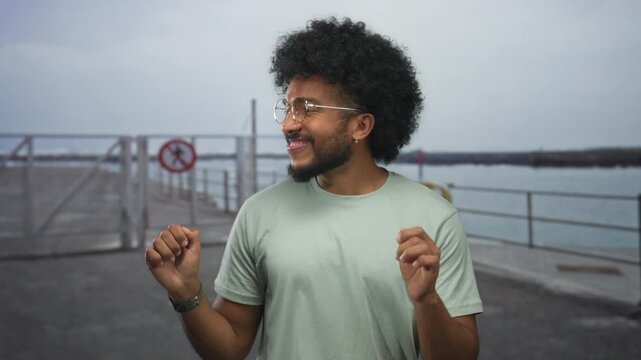 Man dancing joyfully on a seaside promenade with a playful expression, embracing the carefree beach atmosphere, wearing glasses and casual attire, surrounded by the tranquil sea.