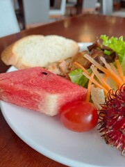Close up of a white plate containing watermelon chunks, fresh salad, cherry tomato, bread, and tropical rambutan