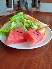 Close up of a white plate containing watermelon chunks, fresh salad, cherry tomato, bread, and tropical rambutan