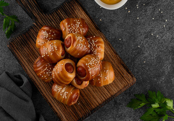 Mini hot dog bites. Sausages baked in a yeast dough cover. Pigs in a blanket on a wooden board on a dark concrete background. Top view.