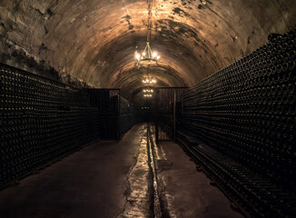 Obraz premium Dark underground wine cellar featuring endless rows of bottles illuminated by soft chandeliers