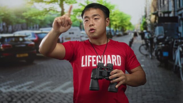 Man wearing red lifeguard shirt holds binoculars and points finger upward on busy street; alertness.