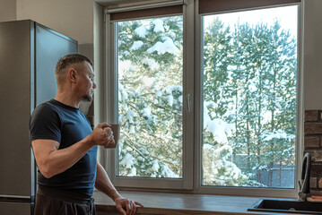 Thoughtful man with mug of hot drink looking out of window to snow scene