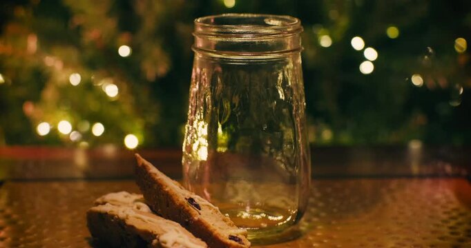 Closeup of Milk Being Poured into Mason Jar with Biscotti Cookies on the side, ready for Santa Claus, Christmas Tree Lights in Cozy Background