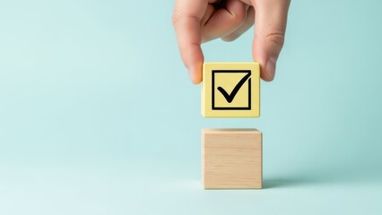 Hand placing a yellow wooden block with a black checkmark symbol on top of another wooden block against a light blue background