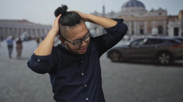 Man with hands on head in vatican building square outdoors, wearing glasses and dark shirt, topknot hairstyle visible; anxiety.