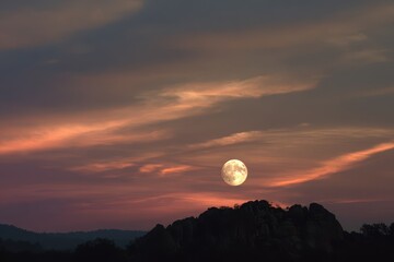 Worm Moon rising behind a silhouetted hill at sunset with dramatic cloudscape