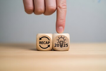 Hand pointing to "RECAP" and "2025" wooden dice with lightbulb icon on wooden table, concept of year summary and future plans.
