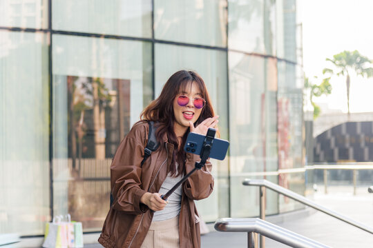 Asian woman enjoys shopping, people embrace fashion lifestyle with shopping bags, vlogging near modern mall, holding phone on stick, posing outside city building
