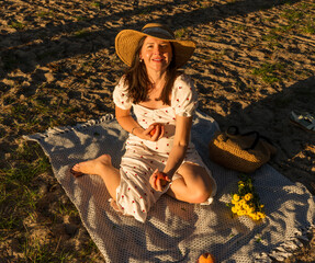 Woman sitting cross legged on a picnic blanket, holding fruit and smiling in warm golden hour light. Natural surroundings and casual pose emphasize joyful summer lifestyle and outdoor relaxation.