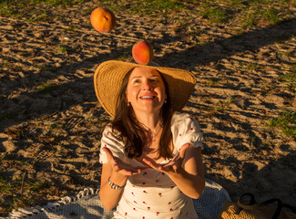Woman playfully tossing ripe peaches while sitting on a picnic blanket during golden hour. Warm light and expressive gesture create a joyful, carefree summer lifestyle scene in a natural outdoor setti