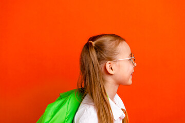 Young girl with glasses smiles in a bright orange background with a green backpack