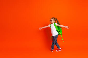 Young girl with green backpack walks joyful on bright orange background showcasing playful school day mood