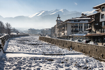A frozen Glazne riverbed with snow-capped boulders creates a textured foreground against a stone wall, buildings, and the Pirin mountain range in Bansko, Bulgaria.