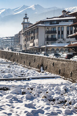 A close-up of a snow-filled Glazne riverbed with a stone retaining wall, showcasing architecture of a ski resort town with mountains in the distance in Bansko, Bulgaria.