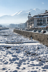 Sunny winter day in the mountain resort of Bansko, Bulgaria. The Glazne riverbed is covered in fresh snow, with traditional buildings lining the street.