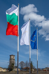 Three flags, including Bulgarian and European Union banners, proudly wave on poles against a bright blue sky with scattered clouds above an old water tower at the train station in Bansko, Bulgaria.